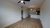 This single-floor space is designed with an open-concept layout that seamlessly connects the living area to the kitchen, creating a bright and airy environment. The living area is finished with light-toned wood-style flooring that extends throughout, lending warmth and continuity to the space. A ceiling fan with an integrated light fixture is centrally mounted, providing both airflow and ambient lighting.

The kitchen is positioned toward the rear of the open floor plan and features a sleek, light-colored countertop island with an integrated sink. A stainless steel refrigerator is visible, complementing the modern aesthetic of the space. Overhead, a combination of pendant lights and track lighting illuminates the kitchen area, adding a contemporary touch.

The neutral white walls provide a clean backdrop throughout, while the open passageways between areas enhance the sense of spaciousness. Additional doorways lead to separate rooms, including what appears to be a utility or laundry area to one side, adding functional versatility. The overall ambiance is modern, clean, and well-lit, making the space feel open and move-in ready.