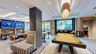 Located at 5919 N Kings Hwy in Alexandria, VA, this single-floor amenity lounge offers a generously sized, open-concept space thoughtfully designed for both social gatherings and leisure activities. The interior showcases a contemporary aesthetic, characterized by large-format stone tile flooring, coffered and tray ceilings with warm recessed and cove lighting, and a combination of structural columns clad in textured dark stone and smooth light wood paneling.

The space is organized into several distinct zones. A gaming area anchors one section, featuring a full-size pool table, a classic arcade cabinet, and a window-facing bar counter with stools that overlook outdoor greenery. A media lounge area includes a built-in wall of large flat-screen televisions framed by brick and wood shelving, accompanied by high-top dining tables and upholstered seating. A separate lounge zone provides comfortable sectional sofas and mixed seating arrangements centered around a striking sculptural pendant light. Additional highlights include a wet bar with open shelving, marble-topped accent tables, and large windows and glass doors that invite natural light and connect the interior to outdoor areas. The overall ambiance is modern, vibrant, and well-appointed.
