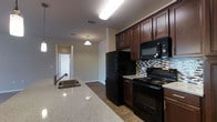 This single-floor residence offers a thoughtfully designed layout with a kitchen that serves as a central highlight. The kitchen is outfitted with rich, dark espresso-toned cabinetry that extends to the ceiling, providing generous storage space. Granite-style countertops add a polished, upscale touch, and a double-basin undermount sink sits along the kitchen island or bar, making meal prep and entertaining convenient. The full suite of black appliances — including a refrigerator, electric range, and over-the-range microwave — creates a cohesive, modern aesthetic. A mosaic tile backsplash in neutral tones adds texture and visual interest behind the range area. The flooring throughout is a warm wood-tone laminate, lending continuity and warmth across the space. A pendant light fixture hangs gracefully over the kitchen area, complementing the overhead fluorescent lighting for ample illumination. The open sightline from the kitchen extends down a hallway toward additional rooms, suggesting a practical and connected floor plan. The overall ambiance is clean, functional, and modern, with a neutral color palette that creates a welcoming and versatile living environment.