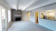This single-floor space offers an open and airy layout anchored by a standout living area with a classic fireplace set against a bold charcoal accent wall. A white mantel adds a clean, contrasting detail, while decorative exposed ceiling beams lend architectural character and a sense of structure to the room. Soft, neutral carpeting covers the living area floor, contributing to a calm and comfortable atmosphere.

The layout opens toward additional functional areas, with a glimpse into a well-organized walk-in storage or pantry space fitted with built-in shelving — a practical and accessible feature. Adjacent to this, the kitchen is partially visible, showing dark countertops, cabinetry, and what appears to be a full-size refrigerator, with hardwood-style flooring transitioning from the carpeted main area.

The walls are painted in a light, neutral grey tone throughout most of the space, complementing the darker accent wall near the fireplace. Ceiling vents and clean white trim throughout suggest a well-maintained interior. Overall, the space presents a balanced blend of functional design and modern, understated aesthetics.