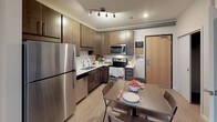 This single-floor unit offers a well-organized layout that balances modern finishes with functional design throughout. The kitchen is a standout feature, outfitted with stainless steel appliances including a full-size refrigerator, electric range, built-in microwave, and dishwasher. Rich dark wood cabinetry contrasts with light countertops, while track lighting illuminates the space. A dining area adjacent to the kitchen provides a comfortable setting for meals.

The living area is compact yet inviting, featuring large windows that allow natural light and urban views to fill the room. Exposed industrial-style ductwork along the ceiling adds a distinctive architectural character to both the living space and bedroom areas.

Two bedrooms offer comfortable accommodations, each featuring sliding-door closets with wire shelving systems for organized storage. Carpeted flooring adds warmth underfoot, and windows provide natural light and city views. One bedroom includes a built-in desk area, making it well-suited for study or work.

Both bathrooms are finished to a high standard, each featuring a glass-enclosed walk-in shower with wood-toned tile surround, a single undermount sink set in a white countertop, dark wood vanity cabinetry, and wood-look flooring, creating a clean, contemporary aesthetic throughout.