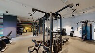 Located at 196 Westfield Rd in Noblesville, Indiana, this single-floor fitness facility offers a comprehensive and thoughtfully designed workout environment spread across a generous footprint. The space is divided into two dedicated exercise rooms, a flexible studio area, and a connecting hallway, providing distinct zones for varied training styles.

The main strength and cardio area is bright and open, featuring large windows that invite natural light and offer outdoor views, creating an energizing atmosphere. This room houses an array of cardio equipment — including treadmills, ellipticals, and stationary bikes — alongside a wall-mounted flat-screen TV for entertainment during workouts. A multi-function cable machine and a full dumbbell rack round out the strength training offerings.

A second exercise room is designed for functional and free-weight training, distinguished by a bold, multicolored accent wall that adds a dynamic visual energy to the space. Dark accent panels frame the room, and the open floor plan accommodates cable systems, weight benches, and ample room for functional movement.

The studio room features light hardwood-style flooring, full-length mirrors along one wall, exposed ductwork overhead, and wall-mounted screens — ideal for group classes, stretching, or guided workouts. Track lighting throughout the facility ensures consistent, well-lit conditions across all areas, while the clean, modern design maintains a professional and motivating ambiance.