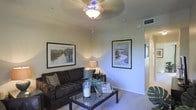 This single-floor residence features a comfortable living room as its central gathering space. The room is finished with neutral-toned walls and plush carpeted flooring, contributing to a warm and inviting atmosphere. A decorative ceiling fan with an ornate light fixture serves as a focal point overhead, providing both ambient illumination and air circulation. Two framed landscape artworks adorn the walls, adding a curated, finished look to the space. The living area accommodates a dark upholstered sofa accented with decorative throw pillows, paired with a sleek glass-top coffee table that holds tasteful decorative accents, including a potted plant and candle arrangements. A small side table with a table lamp adds supplemental lighting and functionality. A hallway visible from the living room suggests additional rooms beyond, with a doorway leading further into the residence. The HVAC vent integrated into the ceiling indicates a climate-controlled environment. Overall, the space presents a clean, well-maintained interior with a cohesive neutral color palette and thoughtful decorative touches throughout.