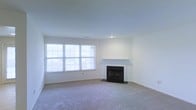 This single-floor residence features a spacious living room that serves as the central focus of the space. The room is finished with neutral wall-to-wall carpeting, which adds warmth and comfort underfoot. The walls are painted a clean, crisp white, creating a bright and open atmosphere throughout.

A standout feature of the living room is the classic fireplace, complete with a simple white mantel and a dark hearth surround, providing a traditional focal point to the space. The fireplace is well-positioned against the wall, making it a natural gathering point for the room.

Natural light is a defining characteristic of this living space, with a large multi-pane window spanning a significant portion of one wall. This window allows an abundance of daylight to flood the room, enhancing the airy, open feel of the interior. The overall design aesthetic is clean and understated, offering a neutral canvas that lends itself well to a variety of interior styles and furnishing choices.