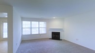 This single-floor residence features a spacious living room that serves as the central focus of the space. The room is finished with neutral wall-to-wall carpeting, which adds warmth and comfort underfoot. The walls are painted a clean, crisp white, creating a bright and open atmosphere throughout.

A standout feature of the living room is the classic fireplace, complete with a simple white mantel and a dark hearth surround, providing a traditional focal point to the space. The fireplace is well-positioned against the wall, making it a natural gathering point for the room.

Natural light is a defining characteristic of this living space, with a large multi-pane window spanning a significant portion of one wall. This window allows an abundance of daylight to flood the room, enhancing the airy, open feel of the interior. The overall design aesthetic is clean and understated, offering a neutral canvas that lends itself well to a variety of interior styles and furnishing choices.