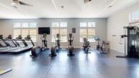 Located at 4202 Forge Cir in Lawrence Township, NJ, this fitness center offers a spacious, single-floor exercise facility designed to support a full range of workout routines. The room is finished with large-format rubber flooring tiles that provide both durability and comfort underfoot, while high ceilings and ceiling fans contribute to an open, well-ventilated atmosphere. Natural light streams in through a row of windows along one wall, brightening the space and offering views of the surrounding exterior. The cardio zone is well-appointed with multiple elliptical trainers, upright and spin bikes, and a bank of treadmills positioned against a bold, motivational mural that energizes the environment with dynamic graphics and fitness-focused messaging. The strength training area features a comprehensive selection of cable machines, multi-function squat racks, weight benches, and a full dumbbell rack, catering to users of all fitness levels. Wall-mounted mirrors expand the sense of space and allow for form monitoring during workouts. A wall-mounted flat-screen TV adds entertainment options for cardio users. The facility also includes a built-in water fountain station near the entrance, ensuring convenience and accessibility. Glass-panel entry doors connect seamlessly to adjacent common areas, completing the clean, modern aesthetic throughout.