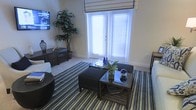 This single-floor space offers a thoughtfully designed interior with a focus on comfort and style. The living room serves as the centerpiece, showcasing large double windows dressed with horizontal blinds and a classic Roman shade that allow abundant natural light to fill the room, creating a warm and airy atmosphere. The soft, neutral wall tones provide a clean backdrop that complements the carefully curated furnishings throughout.

The living area features plush carpeting anchored by a bold blue and gold striped area rug, adding a vibrant touch of color and texture underfoot. Seating is arranged around a glass-top coffee table with a wood X-base frame, accented by decorative blue glass vases and fresh greenery. Dark-stained ottoman seating and additional side chairs provide ample accommodation for guests.

A substantial dark wood media console lines one wall, accompanied by a wall-mounted flat-screen television, making the space equally suited for relaxation and entertainment. A lush decorative indoor tree adds a natural, organic element to the corner of the room, enhancing the overall warmth of the space. The combination of rich wood tones, vibrant accent colors, and generous natural light gives this home a polished yet inviting character.