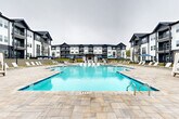Located at 700 Avett Ln in Charlotte, NC, this outdoor amenity space is designed to deliver a resort-style experience within a modern residential community. At the heart of the space is a generously sized swimming pool with crystal-clear blue water, accessible via stainless steel entry rails and surrounded by a broad, stone-paver deck that provides ample room for relaxation and recreation. The pool deck is well-appointed with rows of contemporary white lounge chairs and navy blue market umbrellas, offering shaded seating options throughout the area. A canopied cabana adds an additional touch of resort-style comfort along the poolside. The amenity area is anchored by a single-story clubhouse structure that blends modern farmhouse architectural elements — featuring a combination of white lap siding, dark board-and-batten panels, large multi-pane windows, and a pitched roofline. A covered entry with white columns and glass French doors welcomes visitors into the clubhouse, accessed via a paver walkway framed by manicured lawn. The surrounding multi-story residential buildings feature a cohesive dark and white exterior palette with individual balconies. The entire outdoor space is thoughtfully laid out to encourage community gathering, outdoor leisure, and year-round enjoyment in the Charlotte, NC area.