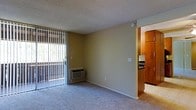 This single-floor residence features a modest, open living area finished with wall-to-wall carpet and neutral-toned walls. A full-height sliding glass door, dressed with vertical blinds, fills the room with natural light and provides direct access to an outdoor balcony, offering views of the surrounding greenery. An in-wall HVAC unit situated beneath the sliding door ensures year-round climate control. The textured ceiling adds a subtle architectural detail consistent throughout the space. Adjacent to the living area, the kitchen is partially visible and features warm wood-toned cabinetry that extends to the upper portion of the walls, providing ample storage. A refrigerator and additional appliances are also noted within the kitchen space. The open transition between the living area and kitchen creates a functional flow, making the layout practical for everyday use. The overall ambiance is simple and utilitarian, with clean lines and minimal ornamentation, lending itself well to personalization.