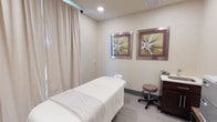 This single-floor treatment room offers a tranquil and professionally appointed space designed for wellness and relaxation services. The room is finished in soft, neutral tones — warm beige walls paired with light grey tile flooring — creating a calming atmosphere conducive to therapeutic treatments.

At the center of the space sits a padded massage table dressed in crisp white linens, positioned to allow easy practitioner access from all sides. Floor-to-ceiling cream drapery lines one wall, adding texture and softness while contributing to the room's sense of privacy and quiet.

Two framed botanical artworks with silver metallic borders are mounted on the wall, featuring muted green and mauve tones that complement the room's overall palette and reinforce its natural, restful aesthetic.

A compact workstation with a dark wood cabinet and laminate countertop is positioned in the corner, accompanied by a cushioned rolling stool — providing a functional area for the practitioner. Recessed ceiling lighting offers gentle, even illumination throughout the space, enhancing the calm and professional ambiance of this well-designed treatment room.