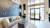 This generously sized single-floor space is designed with a contemporary aesthetic that balances sophistication and comfort. The interior immediately impresses with its soaring vaulted and double-height ceilings, which amplify the sense of openness throughout. Underfoot, large-format herringbone-patterned tile flooring provides a refined, cohesive look across the entire space.

The entrance area features bold navy and teal accent walls adorned with sculptural wall art, creating a striking first impression. Floor-to-ceiling glass doors and expansive windows flood the interior with natural light while providing views of the surrounding exterior. A welcoming lounge zone near the entry includes leather and upholstered seating arranged around a coffee table atop a layered area rug.

Moving further inside, multiple distinct seating arrangements accommodate a variety of uses, from casual lounging to more focused gatherings. A dedicated lounge area features a wall-mounted flat-screen television framed by dark wood shiplap paneling, complemented by velvet accent chairs and bar-height seating. An additional conversation area anchors another zone with a circular metallic coffee table, vibrant area rug, and mixed wood-and-upholstered armchairs. A beverage station and built-in shelving add functional, curated touches. Pendant chandelier lighting and recessed fixtures complete the polished, layered ambiance throughout.
