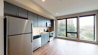 This contemporary single-floor residence showcases a thoughtfully designed open-concept layout that seamlessly connects the kitchen and living areas. The kitchen is a standout feature, outfitted with flat-front slate-blue cabinetry extending to the ceiling for ample storage, a white tile backsplash, and a full suite of stainless steel appliances including a refrigerator, electric range, built-in microwave, and dishwasher. Light wood-tone flooring flows consistently throughout the main living areas, lending warmth and visual continuity to the space.

Large, black-framed floor-to-ceiling windows dominate the living area, inviting generous natural light and offering views of surrounding greenery and the urban skyline beyond. The open living space provides a flexible, airy feel well-suited for a variety of furniture arrangements.

A dedicated walk-in closet offers extensive wire shelving and hanging storage across multiple walls, providing practical and organized storage capacity. The bathroom features the same light wood-tone flooring, clean white walls, contemporary fixtures, and a modern floating-style vanity. Throughout the unit, recessed lighting, crisp white trim, and a neutral color palette contribute to a clean, modern aesthetic that is both functional and inviting.