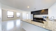 This single-floor space offers a clean, contemporary design centered around an open-concept layout that seamlessly connects the kitchen and living areas. The kitchen is a standout feature, boasting rich dark cabinetry that contrasts elegantly with light-toned granite countertops. A generous island with an embedded electric cooktop provides both functionality and a natural focal point for the space. Stainless steel appliances, including a full-size refrigerator, add a sleek, modern finish. The flooring throughout is a polished terrazzo or stone tile, lending the space a bright, upscale character that reflects natural light effectively. The living area is open and airy, benefiting from multiple windows that allow generous amounts of natural light to fill the room, creating a welcoming and comfortable atmosphere. Recessed ceiling lighting complements the natural light and ensures the space is well-lit at all times. A hallway visible from the main area suggests access to additional rooms or a bathroom, adding to the overall functionality of the layout. The white walls and high ceilings further enhance the sense of openness, making this a versatile and well-appointed single-floor space.