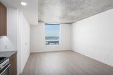 This contemporary single-floor unit showcases a thoughtfully designed open-concept layout that seamlessly connects the kitchen and living area. The space is defined by a striking exposed concrete ceiling, which adds an industrial character and contrasts beautifully with the clean white walls and light wood-tone flooring found throughout. A large window in the living area frames expansive city views, flooding the space with natural light and creating an airy, elevated atmosphere.

The kitchen is well-appointed with warm wood-grain cabinetry extending to the upper walls, a dark subway tile backsplash, light stone countertops, and a full suite of stainless steel appliances including a gas range, built-in microwave, dishwasher, and refrigerator. Recessed lighting provides consistent illumination across the cooking and prep areas.

The bathroom continues the modern aesthetic with large-format light tile flooring, a sleek vanity topped with a gray stone countertop, an illuminated mirror, warm wood cabinetry, and a glass-enclosed shower. The design throughout prioritizes clean lines, neutral tones, and functional details, resulting in a cohesive and polished living environment.