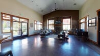 Located at 15720 East 4th Avenue in Spokane Valley, Washington, this single-story community clubhouse offers a thoughtfully designed gathering space with a range of amenities. The centerpiece of the facility is a spacious main lounge featuring soaring vaulted ceilings, polished concrete floors, and an abundance of natural light streaming through large wood-trimmed windows. A dramatic floor-to-ceiling stone fireplace anchors the seating area, which is furnished with leather sofas, accent chairs, and warm, layered décor that creates an inviting atmosphere. A ceiling fan complements the open volume of the room. Adjacent to the main lounge, a well-appointed conference room offers a rustic wood table, leather chairs, a wall-mounted display screen, and a chalkboard — ideal for meetings or collaborative work sessions. The fitness center is well-equipped with elliptical trainers, stationary bikes, a treadmill, free weights, and wall mirrors, all set within a bright, windowed room with rubberized flooring. A kitchenette with wood cabinetry, granite countertops, tile backsplash, and a stainless steel refrigerator provides convenient amenities for gatherings. Warm wood trim details and consistent craftsman-style finishes tie the entire facility together cohesively.