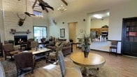 This single-floor space exudes a warm, rustic charm through its thoughtfully designed open-concept layout. The main gathering area commands attention with dramatically vaulted ceilings adorned with exposed wooden beams, creating a sense of grandeur and airiness. Overhead, a combination of a classic chandelier and a ceiling fan complement the elevated roofline, while natural light streams in through large windows, brightening the interior with an inviting glow.

The lounge area is furnished with plush, dark-toned sofas and a variety of accent chairs with nailhead detailing, all arranged around wooden coffee and side tables to encourage comfortable conversation. A decorative round table at the center of the space features an elegant floral arrangement, serving as a natural focal point. Rustic touches, including a mounted deer antler display and area rugs, reinforce the lodge-inspired aesthetic throughout.

Flowing seamlessly from the lounge is a kitchen and bar area, outfitted with warm wood cabinetry, stainless steel appliances, and counter-height seating. This functional space appears well-equipped for hosting and serving guests. The consistent use of hardwood flooring throughout ties the various areas together, contributing to the cohesive, welcoming ambiance of this well-appointed communal environment.
