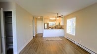 This single-story space is designed with an open, airy layout that promotes a seamless flow between living areas. The living room is a standout feature, showcasing hardwood-style flooring in a warm neutral tone that extends throughout the unit. A fireplace serves as a focal point along one wall, adding both style and function to the space. Sliding glass doors flood the room with natural light while providing direct access to an outdoor area, offering pleasant exterior views.

The kitchen is thoughtfully designed in an open-concept style, featuring a peninsula countertop that connects the cooking area to the main living space. White cabinetry, pendant lighting, and stainless steel appliances give the kitchen a clean, modern aesthetic. A ceiling fan overhead ensures comfortable air circulation. A window beside the kitchen allows additional natural light to brighten the area.

Throughout the space, textured walls, white baseboards, and neutral tones contribute to a cohesive, well-maintained appearance. The overall ambiance is bright, clean, and functional, making it a practical and inviting single-floor living environment.