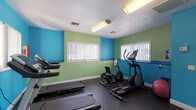 This single-level fitness center offers a dedicated space for cardiovascular and general exercise. The room is immediately recognizable by its bold two-tone wall color scheme — a vivid teal on the lower half and lime green on the upper half — creating an energizing atmosphere conducive to an active workout environment. The flooring throughout is dark rubber matting, providing both durability and cushioning underfoot.

The cardio section is well-appointed with multiple treadmills and an elliptical trainer, ensuring ample availability during peak hours. A stationary recumbent bike and a stability ball round out the aerobic equipment options. A wall-mounted mirror runs along one side of the room, allowing users to monitor their form during exercise. Additional amenities include a cork bulletin board and whiteboard mounted on the wall for informational postings.

Natural light filters in through multiple windows fitted with vertical blinds, keeping the space bright and well-ventilated. Overhead fluorescent lighting supplements illumination throughout. A set of glass double doors opens directly to an outdoor area, where a pool is visible, offering a seamless transition between indoor and outdoor fitness and recreation. The space is also equipped with a fire extinguisher and a security camera, underscoring attention to safety.