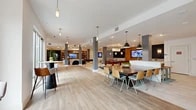 Located at 3701 SW 34th St in Gainesville, FL, this generously sized single-floor community space showcases a contemporary open-plan design with light wood-look tile flooring throughout. The expansive main area seamlessly connects a living room, two dining spaces, and an office, creating a versatile and welcoming environment.

The living room is anchored by a full-wall built-in wood shelving unit housing a large flat-screen TV, complemented by clusters of curved lounge seating, a statement chandelier, and recessed lighting. Adjacent areas feature a game zone with a billiards table, foosball table, and mounted TVs set against warm wood-paneled walls and bold accent lighting.

The two dining areas offer distinct settings — one is a large communal gathering space with long tables and modern pendant lights, while the other features intimate nook-style seating with round white tables, warm wood chairs, soft gray walls, and botanical artwork.

A bright transitional zone with a dramatic floor-to-ceiling gallery wall and gold-accented pendant lighting adds a vibrant artistic focal point. Large glass doors and windows flood the space with natural light and open directly to an outdoor patio, offering views of palm trees. The office provides a quiet, functional retreat completing this well-appointed space.