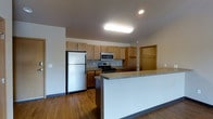 This single-floor space offers a well-designed, open-concept layout that maximizes functionality and flow. The kitchen serves as the centerpiece of the space, featuring warm honey-toned wood cabinetry that provides generous storage with both upper and lower cabinet configurations. Stainless steel appliances include a full-size refrigerator, an electric range, and an over-the-range microwave, delivering a complete and modern cooking setup. The kitchen countertops offer ample workspace, complemented by a peninsula-style breakfast bar with a sleek white finish, ideal for casual dining or additional prep space. Recessed and flush-mounted ceiling lighting ensures bright, even illumination throughout the kitchen area. The flooring throughout is a warm wood-tone laminate that adds a cohesive and inviting aesthetic to the space. Adjacent to the kitchen, a doorway leads to what appears to be a walk-in closet or storage area, visible with a clothing rod, adding practical organizational capacity. The open layout connects seamlessly to the living area, creating a spacious and versatile environment. Neutral wall tones and clean architectural lines contribute to a bright, modern ambiance throughout this well-appointed single-floor space.