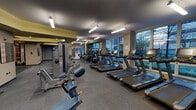 Located at 217 W Washington St in Chicago's Loop neighborhood, this fitness center offers a well-equipped, single-floor facility designed to support a comprehensive workout experience. The space is organized into distinct functional zones, each catering to different training needs.

The cardio area is a standout feature, lined with a row of treadmills and elliptical machines positioned directly in front of large windows. These windows frame views of the surrounding Chicago streetscape and greenery, providing natural light and an engaging backdrop during workouts. The machines are equipped with individual screens for entertainment.

The free weights section includes a full rack of dumbbells in varying sizes, an adjustable bench, and rubber flooring for safety and durability. Strength training machines and cable systems occupy an adjacent area, offering options for targeted muscle training.

Throughout the facility, the design incorporates neutral tones—gray carpeting, yellow accent walls, and a dark ceiling—creating a focused, functional atmosphere. Fluorescent and pendant lighting ensures consistent illumination throughout. Wall-mounted televisions and clearly marked exits contribute to both comfort and safety. The overall layout is open and well-organized, allowing for easy navigation between equipment zones.