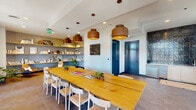 Located at 501 Foster St in downtown Durham, NC, this single-floor communal lounge offers a thoughtfully designed gathering space that blends functionality with contemporary style. The centerpiece of the room is a long, solid-wood communal table surrounded by white chairs, suspended beneath a series of warm-toned drum pendant lights that create an inviting atmosphere for meetings or social gatherings.

Large-format tile flooring extends throughout the space, lending a clean, modern aesthetic. Built-in open shelving along one wall features a textured backdrop and displays curated decorative items, books, and plants, adding character and warmth to the interior. A seating nook with a leather sling chair sits adjacent to the shelving, providing a relaxed reading or lounge area.

On the opposite end of the room, a wet bar area is highlighted by a striking floor-to-ceiling dark glazed tile accent wall, with under-counter refrigeration and cabinetry providing practical amenities. Sheer drape panels frame a window that allows natural light to filter through.

French doors with transom windows open directly onto an outdoor terrace, offering panoramic views of the Durham skyline. A wall-mounted television is positioned nearby, making the space equally suited for presentations or casual viewing. The overall ambiance is modern, curated, and welcoming.