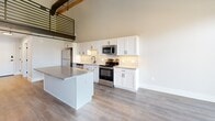 Located at 251 N Rose St in Kalamazoo, MI, this two-story loft blends historic character with modern finishes across two thoughtfully designed floors. The main level hosts an open-concept living room, dining area, and kitchen beneath soaring ceilings that showcase dramatic exposed wooden roof trusses — a striking architectural centerpiece that defines the space's industrial-chic aesthetic. The kitchen is well-appointed with white shaker cabinetry, granite countertops, a subway tile backsplash, a central island, and a full suite of stainless steel appliances including a refrigerator, range, microwave, and dishwasher. Wood-look flooring runs throughout, complementing the warm tones of the timber framework above. A hallway on the main level leads to a full bathroom featuring a tiled tub-surround, granite vanity, white cabinetry, and matte black hardware. A dedicated laundry room with a stacked washer and dryer adds everyday convenience. Upstairs, a steel-railed mezzanine loft overlooks the main living area and leads to the spacious primary bedroom, which features exposed beams and ample room. Two closets provide generous storage. The upper level's open railing and vaulted ceiling create a bright, airy atmosphere throughout.