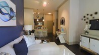This single-floor residence offers a thoughtfully designed open-concept layout that seamlessly connects the living and kitchen areas. The living space is anchored by a plush white sofa accented with patterned throw pillows in navy and white, complemented by a sleek coffee table and a decorative console table adorned with curated accessories. Dark hardwood-style flooring runs throughout, providing a rich contrast to the light-toned walls and bright furnishings.

The kitchen is a standout feature, boasting dark cabinetry with modern pendant lighting suspended above a spacious kitchen island fitted with a granite-style countertop. The island doubles as a breakfast bar with stylish bar stools, making it ideal for casual dining and entertaining. Stainless steel appliances, including an oven range and microwave, add a professional touch to the culinary space. Track lighting overhead ensures the kitchen is well-illuminated.

An entry foyer is visible beyond the kitchen, featuring a paneled door and wall-mounted control panel, providing a welcoming transition into the home. Crown molding along the ceilings adds a refined architectural detail throughout. The overall ambiance is contemporary and chic, blending dark and light tones for a balanced, sophisticated aesthetic.
