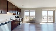 This well-appointed single-floor unit offers a thoughtfully designed open-concept layout that seamlessly connects the kitchen and living area. The kitchen is outfitted with rich, dark-stained cabinetry, light countertops, and a full suite of stainless steel appliances, including a refrigerator, dishwasher, electric range, and over-the-range microwave. The adjoining living area features durable light-toned hardwood-style flooring, neutral walls, and large windows that flood the space with natural light while framing views of the surrounding cityscape and greenery. A sliding glass door leads to a private balcony, extending the living space outdoors. The bedroom is generously sized, finished with plush carpeting, and features a large window with sweeping outdoor views, allowing for excellent natural light. An additional room with hardwood-style flooring and a window offers flexibility as a secondary bedroom or office space. The bathroom is clean and functional, featuring a dark wood vanity with ample storage, light countertops, a large mirror, and hardwood-style flooring. A stacked washer and dryer unit is also visible, adding everyday convenience. Throughout the unit, the consistent use of neutral tones, modern finishes, and natural light creates a cohesive and comfortable living environment.