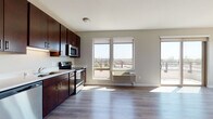 This well-appointed single-floor unit offers a thoughtfully designed open-concept layout that seamlessly connects the kitchen and living area. The kitchen is outfitted with rich, dark-stained cabinetry, light countertops, and a full suite of stainless steel appliances, including a refrigerator, dishwasher, electric range, and over-the-range microwave. The adjoining living area features durable light-toned hardwood-style flooring, neutral walls, and large windows that flood the space with natural light while framing views of the surrounding cityscape and greenery. A sliding glass door leads to a private balcony, extending the living space outdoors. The bedroom is generously sized, finished with plush carpeting, and features a large window with sweeping outdoor views, allowing for excellent natural light. An additional room with hardwood-style flooring and a window offers flexibility as a secondary bedroom or office space. The bathroom is clean and functional, featuring a dark wood vanity with ample storage, light countertops, a large mirror, and hardwood-style flooring. A stacked washer and dryer unit is also visible, adding everyday convenience. Throughout the unit, the consistent use of neutral tones, modern finishes, and natural light creates a cohesive and comfortable living environment.