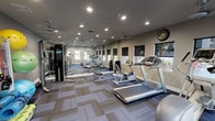 This fitness center is thoughtfully designed to provide a complete workout environment in a single, open-plan space. The room is finished with a patterned modular carpet tile floor and neutral-toned walls, creating a clean and functional atmosphere. Recessed ceiling lighting ensures bright, even illumination throughout, while ceiling fans promote air circulation during workouts.

The cardio section features multiple treadmills, an elliptical trainer, and a stationary bike, each positioned to allow comfortable spacing between users. A dedicated free weights area includes a rack of dumbbells and a flat weight bench, catering to a range of strength training exercises. A multi-station cable machine is also present, offering additional resistance training options.

A full-length mirrored wall runs along one side of the room, visually expanding the space and allowing users to monitor their form during exercise. Several windows fitted with roller shades line the walls, providing natural light and views of the surrounding outdoor greenery while offering the option for privacy. A wall clock keeps users on track during their sessions. The overall layout is spacious, organized, and practical, making this fitness center a well-rounded facility suited to a variety of fitness routines.