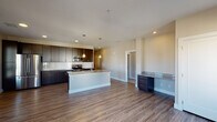 This single-floor unit showcases a contemporary design with a cohesive blend of dark espresso cabinetry, granite countertops, and wood-look flooring that carries throughout the main living areas. The open-concept kitchen is a central highlight, featuring rich dark cabinetry extending to the ceiling, a white subway tile backsplash, stainless steel appliances including a refrigerator, electric range, and microwave, and a central island with granite countertop that provides additional workspace and casual seating. Pendant lighting above the island adds a refined touch. The kitchen flows seamlessly into a spacious living area brightened by multiple windows that allow natural light to fill the room.

The bedroom offers plush carpeting, a ceiling fan, and a generously sized window with exterior views. Two bathrooms serve the unit: a primary bathroom equipped with dark vanity cabinetry, granite countertops, and a glass-enclosed walk-in shower, and a second full bathroom featuring a tub-shower combination and matching vanity finishes. A dedicated laundry area houses full-sized washer and dryer units beneath dark upper cabinetry for added storage. The overall ambiance is clean, modern, and functional.