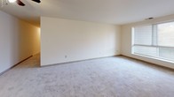 This first-floor apartment at 13516 Greencastle Ridge Road in Burtonsville, Maryland offers a functional and comfortable single-level layout. The living areas are finished with neutral-toned wall-to-wall carpeting and cream-painted walls, creating a clean and versatile backdrop throughout. Natural light filters in through slatted blinds on multiple windows, brightening the generously sized carpeted rooms. Several of the bedrooms or flexible-use rooms feature ample floor space, with at least one room equipped with a ceiling fan and a closet. The galley-style kitchen is well-appointed with warm oak cabinetry lining both sides, light stone-look countertops, tile flooring, and white appliances including a range and refrigerator. A louvered door at the end of the kitchen leads to additional storage or utility space. The unit includes two bathrooms, each featuring tile flooring, a wood-toned vanity with a stone-look composite countertop, an integrated sink, a large mirror, and a towel bar. The consistent finishes throughout the bathrooms provide a cohesive look. The overall floor plan, as seen in the dollhouse view, reflects an efficient and well-organized layout suited for everyday living.
