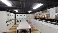 This laundromat offers a practical and efficiently organized space designed to accommodate a high volume of laundry needs. The layout features a generous row of top-loading washing machines along one side and a combination of stacked and front-loading washers and dryers along the opposite wall, providing ample capacity for customers. At the center of the room, a long, white folding table offers plenty of surface area for sorting and folding laundry with ease. The flooring features a distinctive checkered pattern in warm tones, adding a touch of visual interest to the utilitarian space. Overhead, an industrial-style exposed ceiling with dark-painted beams and pipes is illuminated by bright fluorescent strip lighting, ensuring the room is well-lit throughout. A standout decorative element is the large, colorful mural covering one wall, depicting a lively streetscape scene that adds warmth and character to the environment. Storage lockers or utility panels are also visible along the back wall, contributing to the space's functional design. Overall, the laundromat balances practicality with a welcoming atmosphere, making it a comfortable and fully equipped destination for all laundry needs.