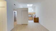 This single-floor space offers a clean, minimalist design characterized by crisp white walls and light-toned flooring that together create a bright and airy atmosphere throughout. The open living area is a standout feature, bathed in natural light streaming through a overhead skylight that draws the eye upward and emphasizes the generous ceiling height. A spherical pendant light complements the skylight, adding a subtle design touch to the space. Adjacent to the living area, the kitchen is efficiently laid out with light natural wood cabinetry, a white refrigerator, and a sink set beneath the upper cabinets. A dark wood-toned breakfast bar or counter provides a visual contrast and serves as a practical divider between the kitchen and the main living space. A built-in closet or partition wall is centrally positioned, helping to define distinct zones within the open floor plan. A hallway visible toward the rear of the space leads to additional rooms, suggesting a thoughtful and functional layout. The overall ambiance is clean, neutral, and well-lit, making the space feel open and versatile.