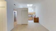 This single-floor space offers a clean, minimalist design characterized by crisp white walls and light-toned flooring that together create a bright and airy atmosphere throughout. The open living area is a standout feature, bathed in natural light streaming through a overhead skylight that draws the eye upward and emphasizes the generous ceiling height. A spherical pendant light complements the skylight, adding a subtle design touch to the space. Adjacent to the living area, the kitchen is efficiently laid out with light natural wood cabinetry, a white refrigerator, and a sink set beneath the upper cabinets. A dark wood-toned breakfast bar or counter provides a visual contrast and serves as a practical divider between the kitchen and the main living space. A built-in closet or partition wall is centrally positioned, helping to define distinct zones within the open floor plan. A hallway visible toward the rear of the space leads to additional rooms, suggesting a thoughtful and functional layout. The overall ambiance is clean, neutral, and well-lit, making the space feel open and versatile.