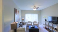 This single-floor living space offers a clean, comfortable layout centered around a well-appointed living room. The room is finished with light-toned walls that contribute to an open, airy atmosphere, complemented by soft carpeted flooring throughout. A ceiling fan with an integrated light fixture serves as a functional focal point overhead, providing both lighting and air circulation.

Natural light flows generously into the space through a large sliding glass door fitted with vertical blinds, which also provides access to an outdoor area. The seating arrangement includes a plush sofa adorned with decorative pillows, two matching armchairs, and a bold navy upholstered ottoman that doubles as a coffee table, adding a contemporary contrast to the neutral surroundings.

A large abstract artwork on the left wall introduces color and visual interest, while a console table on the right holds a decorative plant, adding a touch of greenery. The overall ambiance is calm and inviting, with a balanced mix of comfort and style that makes the living area feel both functional and welcoming.