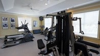 Located at 901 Falls Park Dr in Sanford, NC, this fitness center offers a well-equipped, single-floor workout facility designed to support a range of exercise routines. The space is brightened by multiple windows fitted with white blinds, allowing ample natural light to fill the room while maintaining privacy. Ceiling fans provide steady air circulation throughout, contributing to a comfortable environment during workouts. The fitness center is outfitted with a multi-station cable strength training machine, an elliptical trainer, a stationary upright bike, and additional cardio equipment, giving users access to both cardiovascular and resistance training options. A wall-mounted television is positioned for easy viewing during exercise. Recessed lighting supplements the natural light, ensuring the space remains well-lit at all hours. Crown molding and a tray ceiling detail lend a polished, finished look to the interior. The flooring is carpeted, adding a degree of comfort underfoot. A posted rules board is visible on the wall, indicating the space is a shared community amenity. Overall, the fitness center is a practical and thoughtfully appointed space for residents seeking convenient access to exercise facilities.