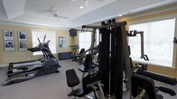Located at 901 Falls Park Dr in Sanford, NC, this fitness center offers a well-equipped, single-floor workout facility designed to support a range of exercise routines. The space is brightened by multiple windows fitted with white blinds, allowing ample natural light to fill the room while maintaining privacy. Ceiling fans provide steady air circulation throughout, contributing to a comfortable environment during workouts. The fitness center is outfitted with a multi-station cable strength training machine, an elliptical trainer, a stationary upright bike, and additional cardio equipment, giving users access to both cardiovascular and resistance training options. A wall-mounted television is positioned for easy viewing during exercise. Recessed lighting supplements the natural light, ensuring the space remains well-lit at all hours. Crown molding and a tray ceiling detail lend a polished, finished look to the interior. The flooring is carpeted, adding a degree of comfort underfoot. A posted rules board is visible on the wall, indicating the space is a shared community amenity. Overall, the fitness center is a practical and thoughtfully appointed space for residents seeking convenient access to exercise facilities.
