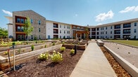Located at 300 Commerce Ave in LaGrange, Georgia, this thoughtfully designed residential community blends contemporary architecture with inviting outdoor living spaces. The multi-building development features a striking combination of brick, board-and-batten siding, and warm wood-toned exterior cladding accented by bold red window surrounds and private balconies on upper levels.

The heart of the community is a beautifully landscaped courtyard that serves as a central gathering hub. Raised wooden garden beds line concrete walkways, planted with a variety of flowering shrubs and seasonal greenery, lending the space a charming, agrarian character. A substantial timber pergola anchors an outdoor kitchen and grilling area, providing a shaded retreat ideal for social gatherings.

Recreational amenities are a standout feature of this property. A well-manicured putting green offers a leisurely activity just steps from residential buildings, while a dedicated bocce ball court with paved brick surrounds adds to the community's engaging outdoor experience. Open lawn areas provide additional green space throughout the grounds.

Perimeter fencing with a ranch-style wood design gives the community a welcoming, neighborhood feel, while mature trees and curated landscaping throughout ensure a lush, well-maintained environment. The overall ambiance is one of active, community-centered living set within a clean and modern architectural framework.