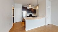 This single-floor residence offers a thoughtfully designed layout with a clean, modern aesthetic throughout. The open-concept kitchen serves as the heart of the space, showcasing rich dark cabinetry with brushed nickel hardware, a white quartz curved peninsula with an integrated sink, and a full suite of stainless steel appliances including a refrigerator, electric range, and built-in microwave. A tile backsplash and stylish pendant lighting overhead add polished detail to the cooking area. The living area is bright and airy, featuring warm hardwood flooring, neutral white walls, and a ceiling fan that enhances comfort and airflow. Natural light filters in through window treatments, creating a welcoming atmosphere. A doorway connects the living space to the carpeted bedroom areas, which offer a quieter, softer underfoot experience. The bedrooms are well-proportioned with ceiling fans, and sliding doors with vertical blinds allow for natural light and privacy. The bathroom is finished with tile flooring, a full soaking tub with a tiled surround, a dark-framed vanity mirror, and a white countertop with ample storage below. Additional utility space with extra cabinetry and countertop surfaces provides practical convenience throughout the home.