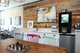 Located at 6860 Bebout Dr in Norcross, Georgia, this single-floor community clubhouse is a thoughtfully designed gathering space that blends rustic and contemporary elements throughout. The centerpiece of the main lounge is a dramatic floor-to-ceiling stacked stone fireplace accented by reclaimed wood wall paneling, anchoring a generously proportioned seating area beneath soaring vaulted ceilings with exposed wooden beams. A statement chandelier with industrial-style pendant lights adds a warm, inviting glow to the space. Hardwood-style flooring runs throughout, complementing the eclectic furnishings that include leather armchairs, a plush sectional, and bold, colorful accent pillows that bring energy and personality to the room. Large floor-to-ceiling glass doors and expansive windows flood the interior with abundant natural light while offering unobstructed views of the surrounding landscaped grounds. A dedicated coffee bar area features a Starbucks station with pristine white quartz countertops, a reclaimed wood accent wall, and a sink — creating a welcoming café-style nook. A work and game area provides additional functionality for residents. The overall ambiance is modern-casual with rustic warmth, making this clubhouse a versatile and inviting communal hub.