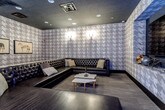 This single-floor lounge is defined by its cohesive, club-inspired aesthetic, anchored by floor-to-ceiling houndstooth-patterned wallcovering in muted tones of grey and taupe that wraps continuously around the entire room. The dark ceiling, outfitted with recessed can lighting, reinforces the intimate, moody ambiance, while decorative wall sconces with warm candlelight-style bulbs provide accent illumination at key points throughout the space.

The primary seating area features a large L-shaped sectional sofa in dark tufted leather, arranged around a pair of white marble-topped coffee tables. Framed equestrian artwork adorns one wall, adding a refined, editorial touch. A large flat-screen television is mounted within a dark recessed frame on one wall, creating a clean, built-in appearance suited for entertainment or presentations.

A bar counter extends along one side of the room, accompanied by a row of leather-topped industrial-style bar stools, making the space well-suited for social gatherings. An additional open area features a bench-style ottoman and open floor space, with a fitness or activity ball visible, suggesting multi-purpose functionality. Dark hardwood-style flooring runs throughout, completing the upscale, lounge-forward character of this versatile space.