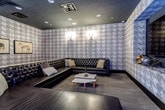 This single-floor lounge is defined by its cohesive, club-inspired aesthetic, anchored by floor-to-ceiling houndstooth-patterned wallcovering in muted tones of grey and taupe that wraps continuously around the entire room. The dark ceiling, outfitted with recessed can lighting, reinforces the intimate, moody ambiance, while decorative wall sconces with warm candlelight-style bulbs provide accent illumination at key points throughout the space.

The primary seating area features a large L-shaped sectional sofa in dark tufted leather, arranged around a pair of white marble-topped coffee tables. Framed equestrian artwork adorns one wall, adding a refined, editorial touch. A large flat-screen television is mounted within a dark recessed frame on one wall, creating a clean, built-in appearance suited for entertainment or presentations.

A bar counter extends along one side of the room, accompanied by a row of leather-topped industrial-style bar stools, making the space well-suited for social gatherings. An additional open area features a bench-style ottoman and open floor space, with a fitness or activity ball visible, suggesting multi-purpose functionality. Dark hardwood-style flooring runs throughout, completing the upscale, lounge-forward character of this versatile space.