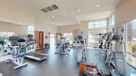 This fitness center in Baton Rouge, Louisiana, is a thoughtfully designed, single-floor facility that balances functionality with an open, inviting atmosphere. The space is characterized by soaring ceilings and an abundance of large, floor-to-ceiling windows along multiple walls, flooding the interior with natural light and offering views of the surrounding landscape.

The main workout area is generously proportioned, providing ample room for a diverse selection of equipment. Cardio machines include treadmills, ellipticals, and a rowing machine, while a comprehensive range of strength training equipment—including cable machines, weight benches, and a full dumbbell rack—supports varied fitness routines. Dark, durable rubber flooring covers the workout area, offering both safety and practicality.

Strategic placement of large wall mirrors enhances the sense of space while allowing users to monitor their form. Recessed lighting supplements the natural light throughout the room. A separate hallway leads to additional amenity spaces, including restrooms with warm wood-toned doors. The entry features a glass-and-wood door, a hand sanitizer station, and posted fitness center guidelines, contributing to a clean, well-organized environment overall.