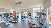 This fitness center in Baton Rouge, Louisiana, is a thoughtfully designed, single-floor facility that balances functionality with an open, inviting atmosphere. The space is characterized by soaring ceilings and an abundance of large, floor-to-ceiling windows along multiple walls, flooding the interior with natural light and offering views of the surrounding landscape.

The main workout area is generously proportioned, providing ample room for a diverse selection of equipment. Cardio machines include treadmills, ellipticals, and a rowing machine, while a comprehensive range of strength training equipment—including cable machines, weight benches, and a full dumbbell rack—supports varied fitness routines. Dark, durable rubber flooring covers the workout area, offering both safety and practicality.

Strategic placement of large wall mirrors enhances the sense of space while allowing users to monitor their form. Recessed lighting supplements the natural light throughout the room. A separate hallway leads to additional amenity spaces, including restrooms with warm wood-toned doors. The entry features a glass-and-wood door, a hand sanitizer station, and posted fitness center guidelines, contributing to a clean, well-organized environment overall.