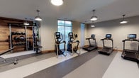 This modern fitness center offers a well-appointed workout environment designed to support a range of exercise routines. The space features a clean, functional layout with durable striped rubber flooring that provides both safety and comfort underfoot. Overhead, a series of industrial-style pendant lights deliver consistent, warm illumination throughout the room, complementing the natural light that filters in through large windows along one wall.

The cardio section is a highlight of the facility, equipped with multiple elliptical cross-trainers and treadmills, each featuring integrated digital display screens for tracking workout metrics. Along one wall, a dedicated strength training and functional fitness area provides cable machines and resistance equipment, offering users a comprehensive full-body workout experience.

The neutral color palette of the walls and the open layout contribute to a spacious, uncluttered atmosphere that is both inviting and practical. The combination of natural light, ample equipment variety, and thoughtful design makes this fitness center a well-rounded amenity suited for both casual exercisers and dedicated fitness enthusiasts alike.
