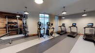 This modern fitness center offers a well-appointed workout environment designed to support a range of exercise routines. The space features a clean, functional layout with durable striped rubber flooring that provides both safety and comfort underfoot. Overhead, a series of industrial-style pendant lights deliver consistent, warm illumination throughout the room, complementing the natural light that filters in through large windows along one wall.

The cardio section is a highlight of the facility, equipped with multiple elliptical cross-trainers and treadmills, each featuring integrated digital display screens for tracking workout metrics. Along one wall, a dedicated strength training and functional fitness area provides cable machines and resistance equipment, offering users a comprehensive full-body workout experience.

The neutral color palette of the walls and the open layout contribute to a spacious, uncluttered atmosphere that is both inviting and practical. The combination of natural light, ample equipment variety, and thoughtful design makes this fitness center a well-rounded amenity suited for both casual exercisers and dedicated fitness enthusiasts alike.