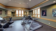 This fitness center is thoughtfully designed to provide a comfortable and functional workout environment. The space is characterized by sage green walls accented with warm wood trim, creating an inviting and cohesive aesthetic throughout. Vaulted ceilings in the main workout area add a sense of openness, complemented by a combination of recessed lighting and adjustable track lighting that ensures the space is well-lit at all times.

The cardio area is generously equipped with Matrix treadmills, elliptical trainers, upright and recumbent stationary bikes, and additional aerobic machines to accommodate a variety of fitness routines. Wall-mounted flat-screen televisions are positioned for easy viewing during workouts. A dedicated stretching and floor exercise area features a large wall mirror that runs the length of the room, enhancing the sense of space and supporting form-focused training.

Multiple wood-framed windows line the walls, allowing natural light to filter in and offering views of the surrounding greenery. Rubber flooring throughout provides safety and durability underfoot. A built-in bench with lower cubbies and wall-mounted coat hooks offers a convenient changing and storage area. Sports-themed artwork displayed on the walls adds an energetic and motivational touch to the overall ambiance.