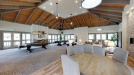 This impressive single-floor gathering space is defined by its dramatic vaulted ceiling, where warm wood planking and exposed gray structural beams draw the eye upward and lend a lodge-inspired character to the interior. Large ceiling fans and carefully placed pendant and chandelier lighting ensure the space is well-lit and comfortable throughout. Abundant windows lining the perimeter walls flood the interior with natural light and provide views to the surrounding exterior.

The open floor plan is thoughtfully organized into distinct functional zones. A recreational area features a full-size pool table and a shuffleboard table, making it well-suited for social gatherings and entertainment. Adjacent to this, a comfortable lounge area is furnished with plush upholstered seating arranged to encourage conversation and relaxation. A dining area with a round wood table and upholstered chairs completes the layout, offering a space for more formal seated gatherings.

The flooring transitions between hardwood and carpet, subtly delineating each area while maintaining a cohesive, modern-rustic aesthetic. The combination of high ceilings, warm wood tones, neutral furnishings, and generous natural light creates a welcoming and versatile atmosphere well-suited for a wide range of social activities.